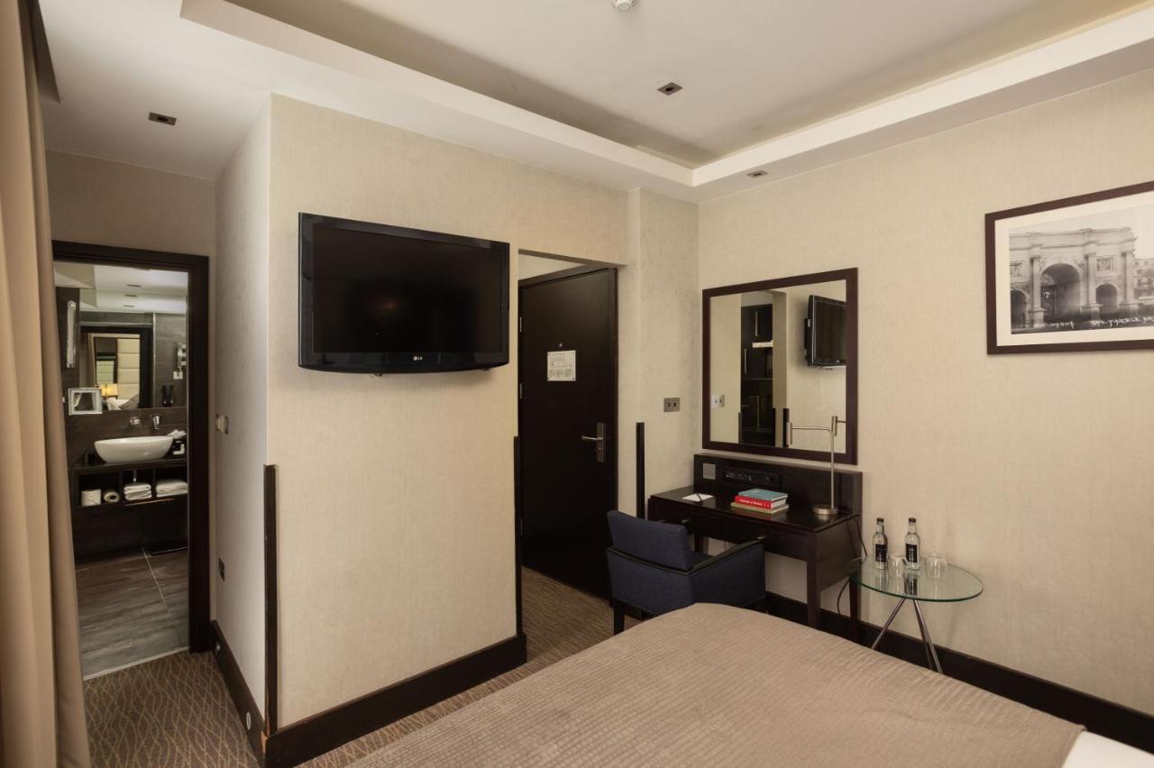 Montcalm Brewery Townhouse Londres Exterior foto The photo shows a hotel room featuring a double bed with a neutral-colored bedspread. On one wall, there is a flat-screen TV mounted. Adjacent to the bed, there is a stylish desk with a chair and a small glass table. A mirror is positioned above the 