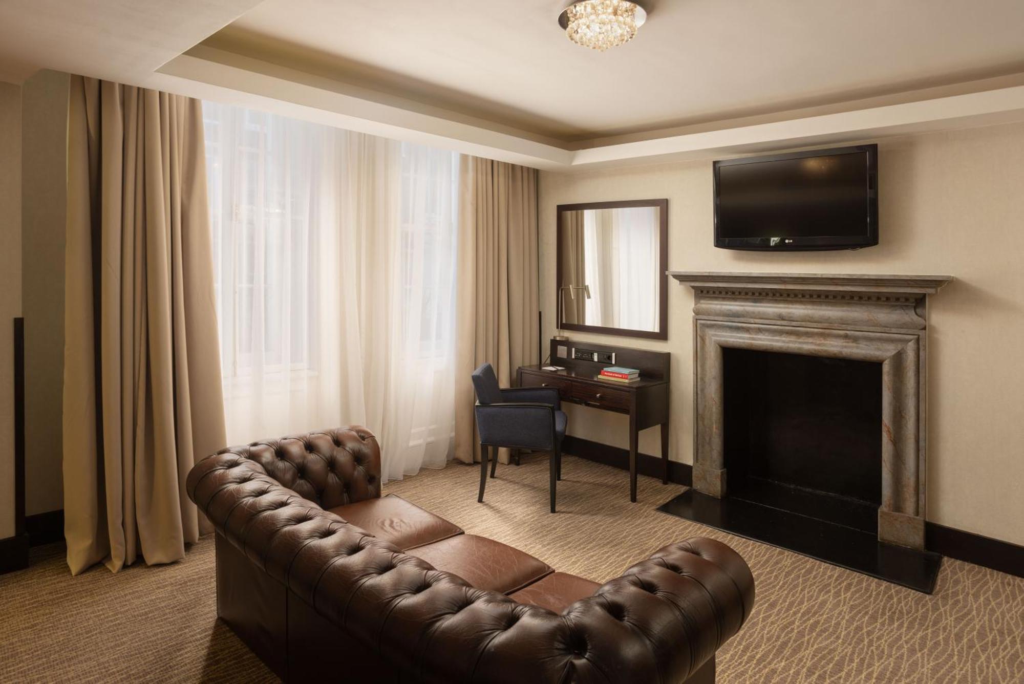 Montcalm Brewery Townhouse Londres Exterior foto The photo shows a stylish interior of a room, likely a living area or a hotel suite. It features a dark leather chesterfield sofa with tufted upholstery. Opposite the sofa is a small wooden desk with a chair, positioned near a wall-mounted flat-scree