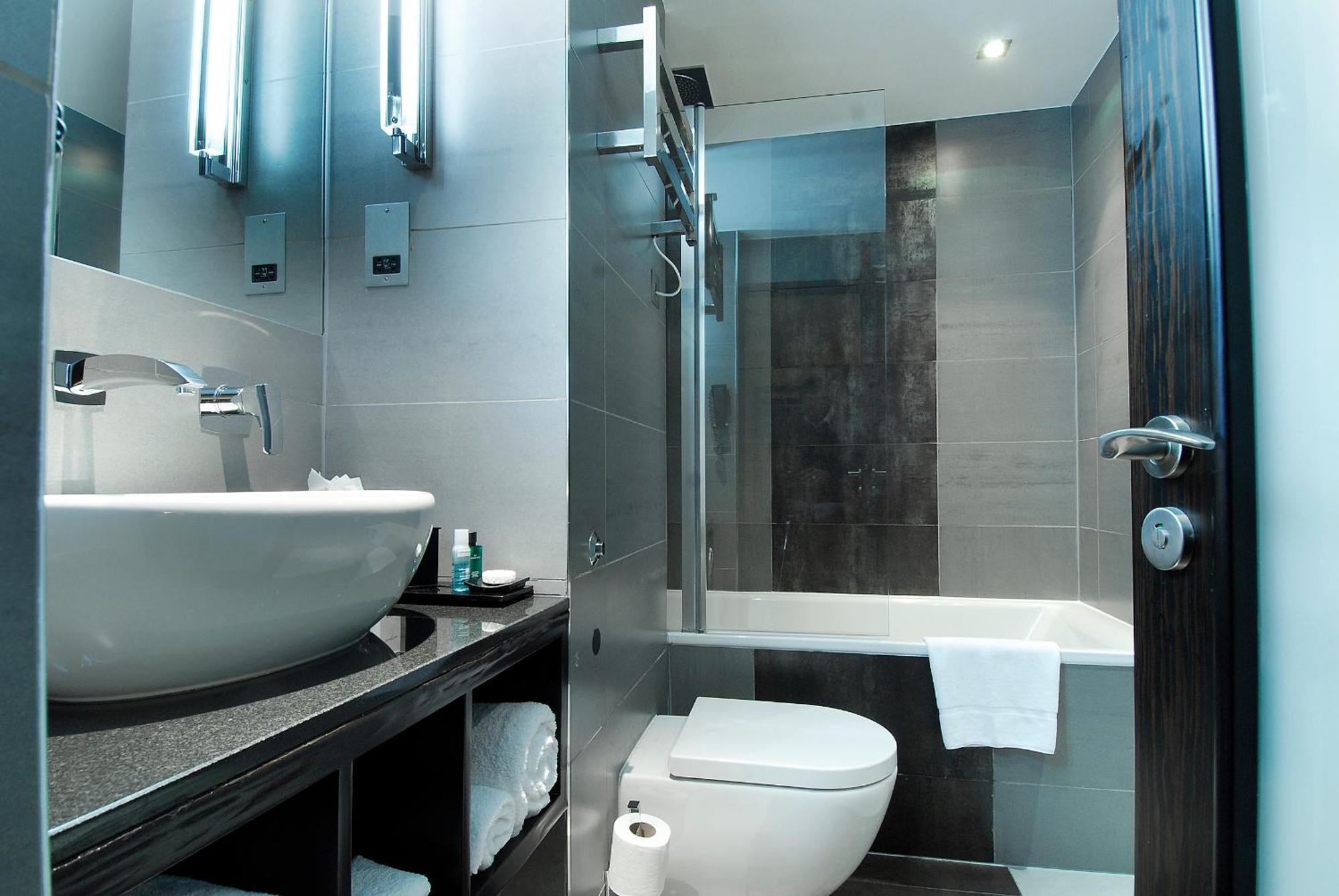 Montcalm Brewery Townhouse Londres Exterior foto The photo shows a modern bathroom with sleek design elements. It features a curved white sink on a dark countertop, with a mirror above that is illuminated by stylish lighting fixtures. Next to the sink, there's a toilet, and on the opposite side, a 