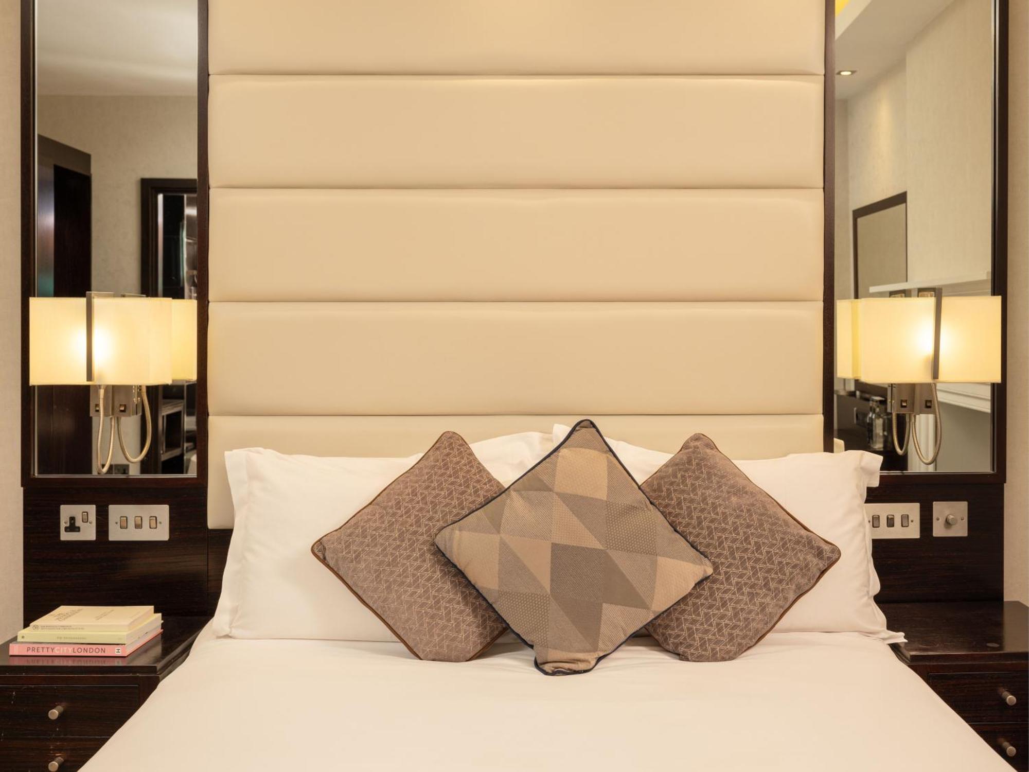 Montcalm Brewery Townhouse Londres Exterior foto The image shows a cozy bedroom setting. The bed features a headboard upholstered in a light beige fabric. It is adorned with three decorative pillows in varying patterns and colors, placed on white bedding. On either side of the bed, there are modern