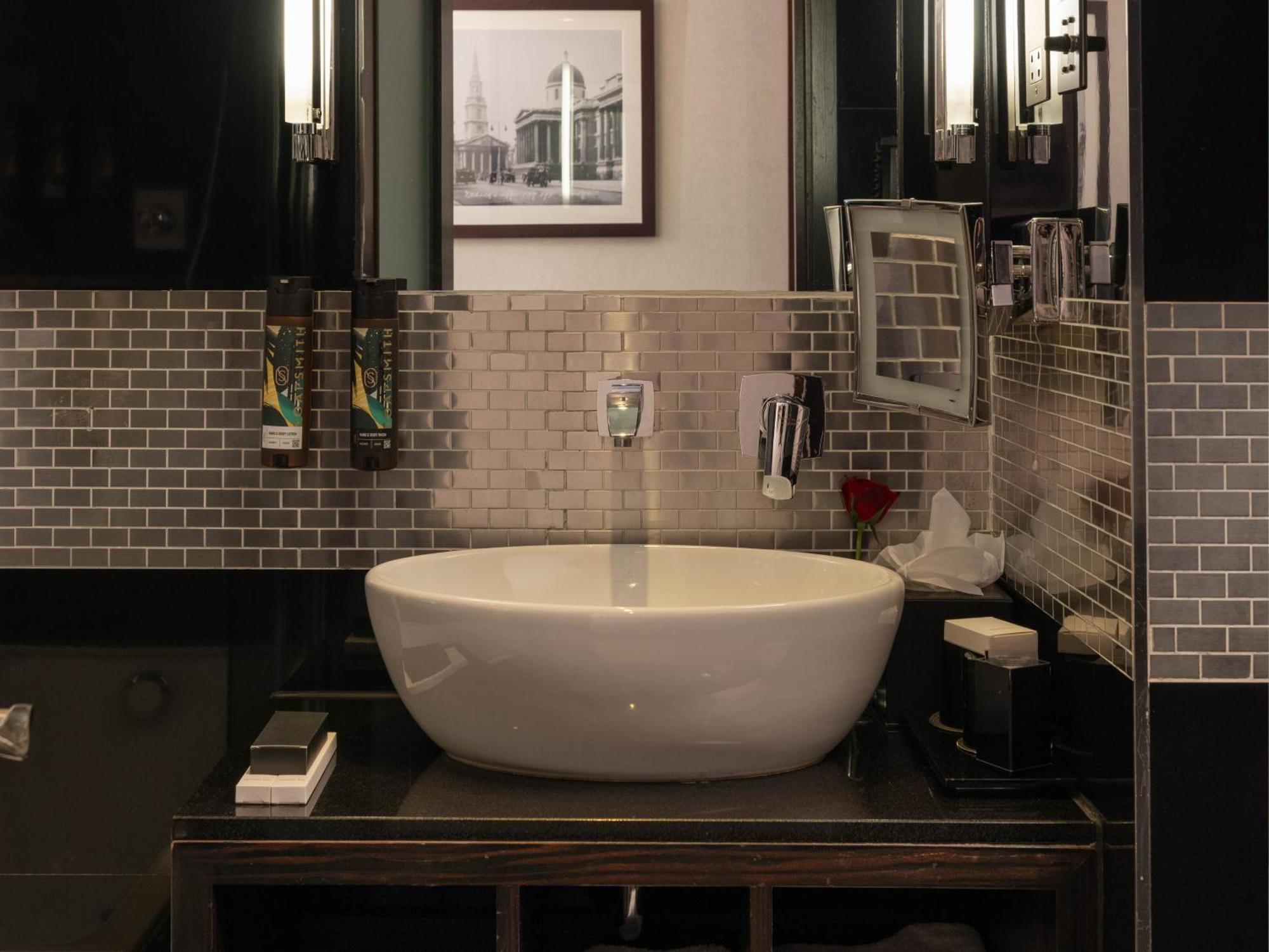 Montcalm Brewery Townhouse Londres Exterior foto The photo shows a modern bathroom with a stylish design. It features a large, round white sink placed on a dark wooden vanity. The walls are adorned with a mix of gray and silver tile, creating a sleek and contemporary look. Above the sink, there is 