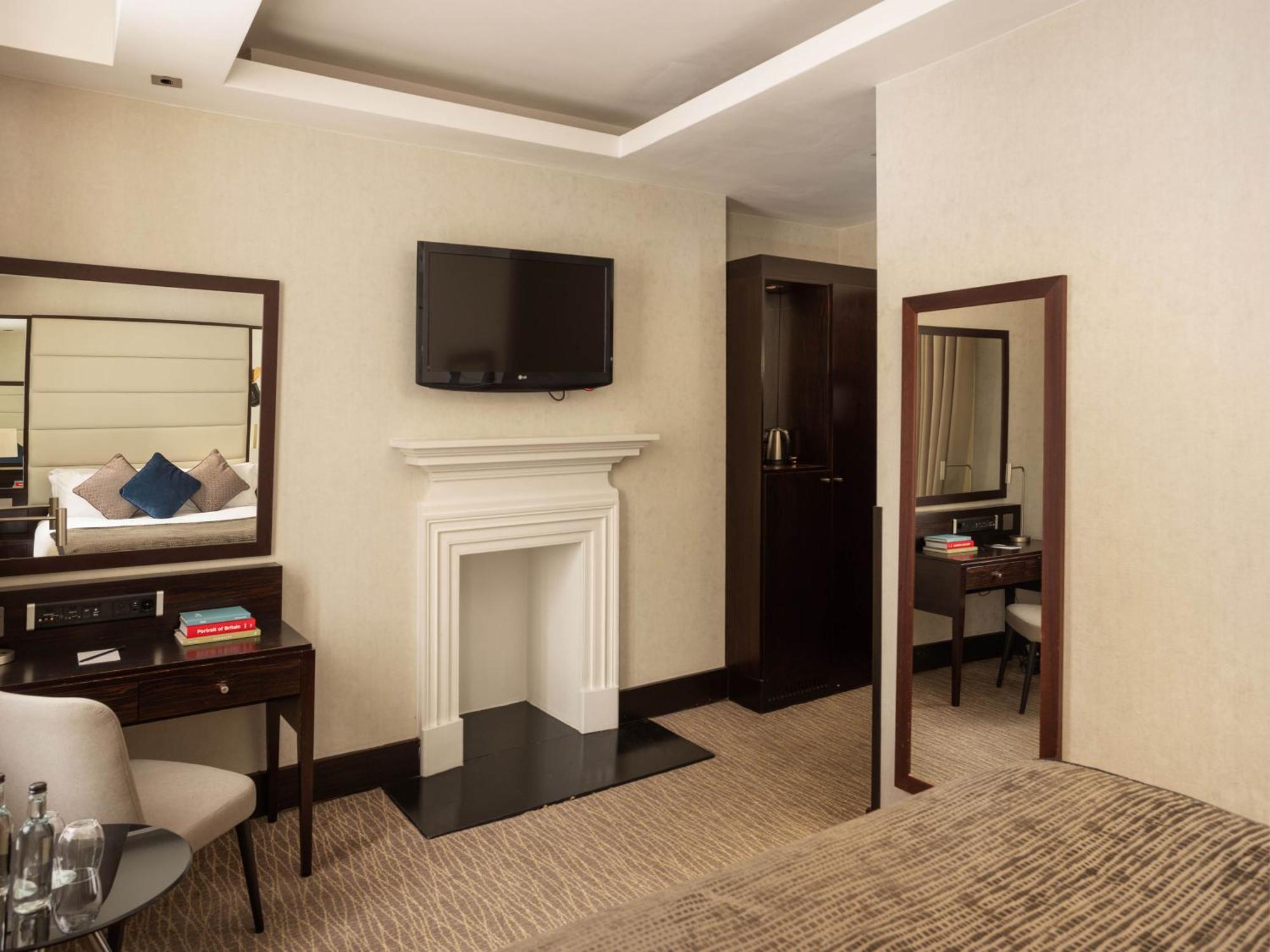 Montcalm Brewery Townhouse Londres Exterior foto The photo depicts a stylish hotel room. It features a double bed, a large flat-screen TV mounted on the wall, and a decorative fireplace. There is a wooden desk with a chair beside it, as well as a mirror hanging on the wall. The decor is modern and 