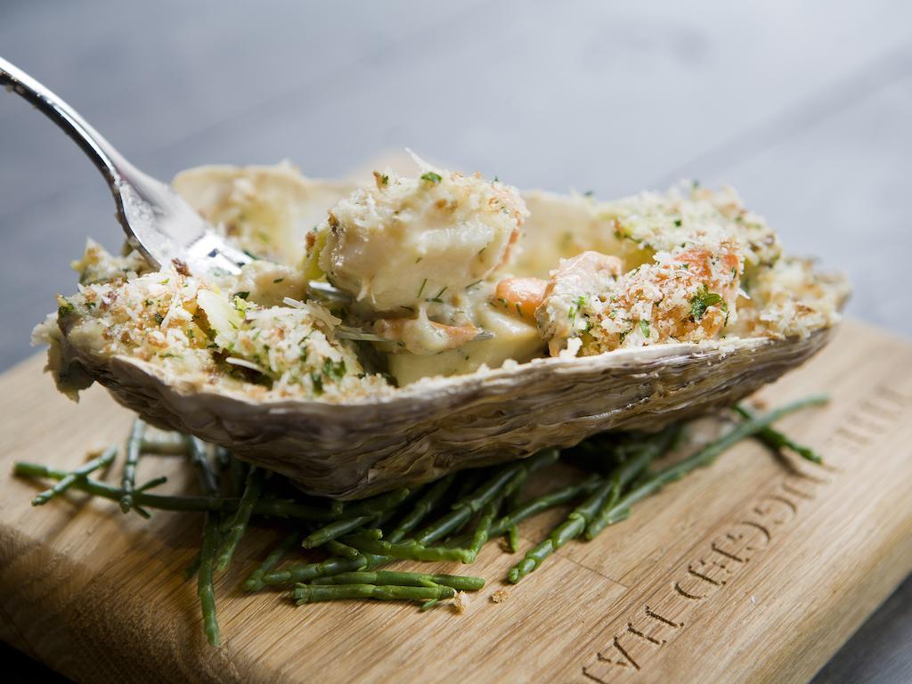 Montcalm Brewery Townhouse Londres Exterior foto The photo shows a large oyster shell filled with a mixture that appears to include seafood, herbs, and breadcrumbs. The dish is presented on a wooden board with some greenery, possibly a type of seaweed or garnishing. A fork is placed beside the oyst