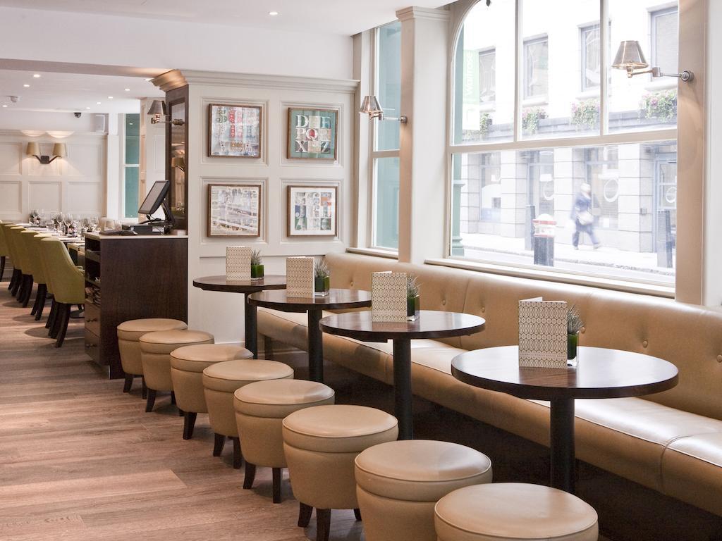 Montcalm Brewery Townhouse Londres Exterior foto The photo displays a modern café or restaurant interior. There is a cozy seating area featuring light-colored, upholstered benches along the wall, accompanied by small round tables. The tables are paired with circular stools. The decor includes frame