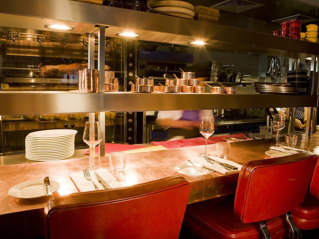 Montcalm Brewery Townhouse Londres Exterior foto The photo shows a restaurant interior featuring a bar or counter area. There are red chairs arranged around a copper-topped table where dining utensils and plates are set. Above the counter, there are shelves displaying various cooking pots and kitch