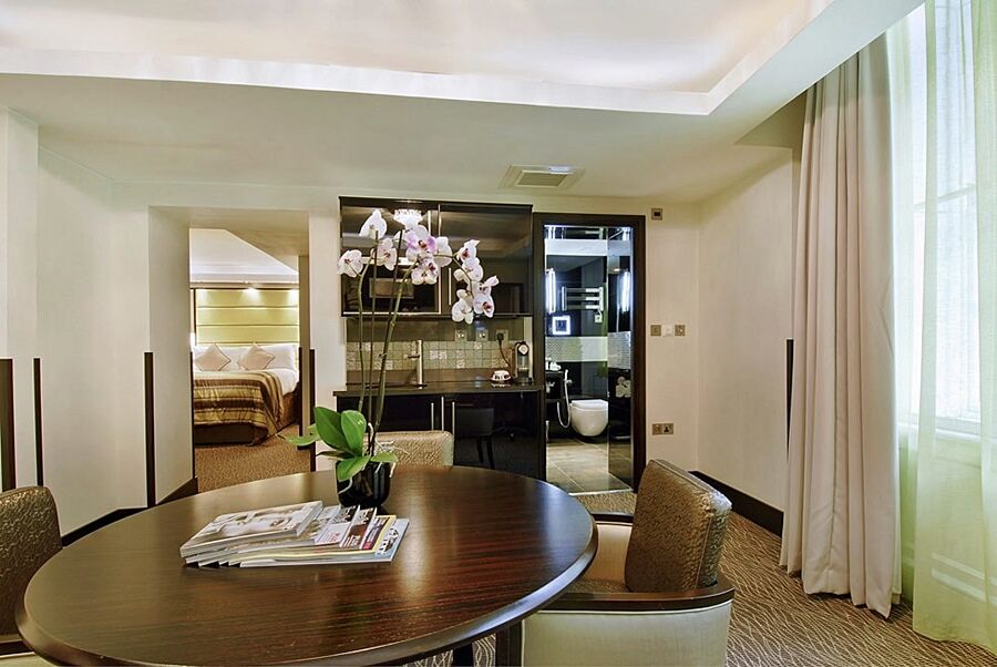 Montcalm Brewery Townhouse Londres Exterior foto The photo shows a stylish hotel room or suite. In the foreground, there is a round dining table with a few magazines on it. Surrounding the table are upholstered chairs. To the right, there are large windows with light-colored curtains, allowing natu