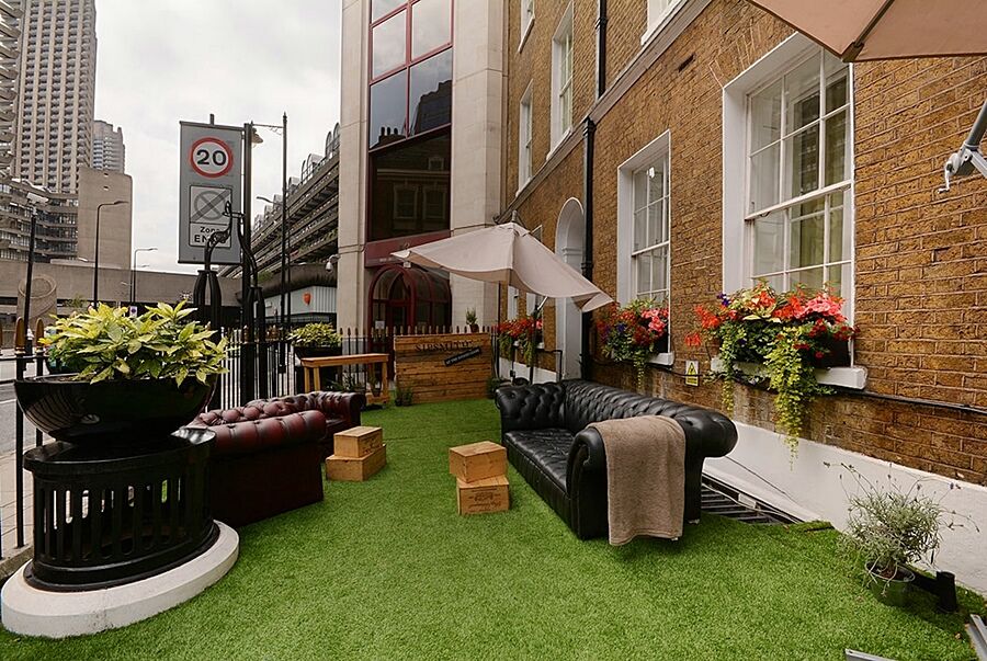 Montcalm Brewery Townhouse Londres Exterior foto The photo depicts a charming outdoor space, likely a patio or garden area. It features two comfortable-looking leather sofas, one of which is a deep burgundy color, placed on a green artificial lawn. There are wooden crates that could serve as side t